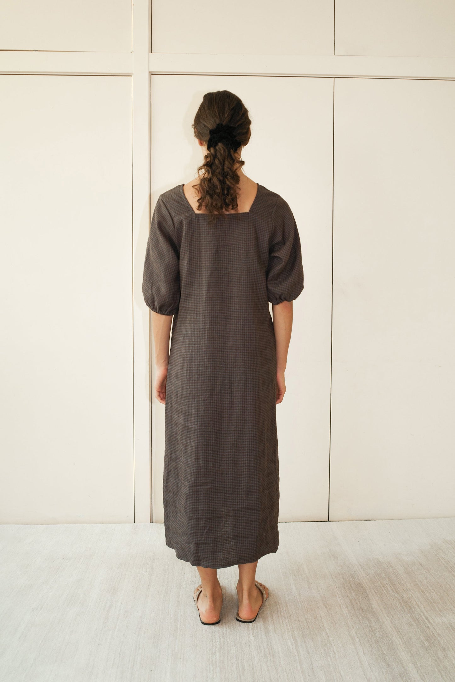 A barefoot person with curly hair in a low ponytail faces away from the camera, wearing Deiji Studios Squared Dress - Slate Check, a loose-fitting organic linen dress. The backdrop is a plain, light-colored wall.