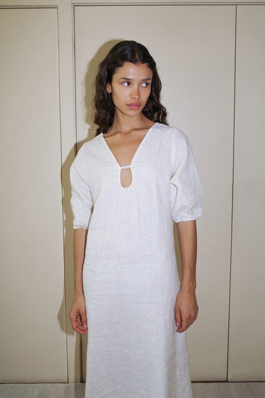 A woman with wavy brown hair wears the Squared Dress, a light-colored long dress with elbow-length sleeves and a small keyhole neckline, standing by beige double doors and glancing to the side.