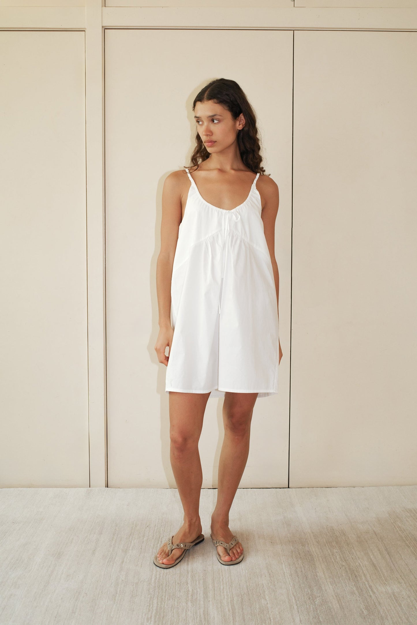 A woman in the Double V Dress - White, a loose organic cotton everyday dress, wears sandals and stands on a light carpet before cream paneled doors, gazing slightly to the side.