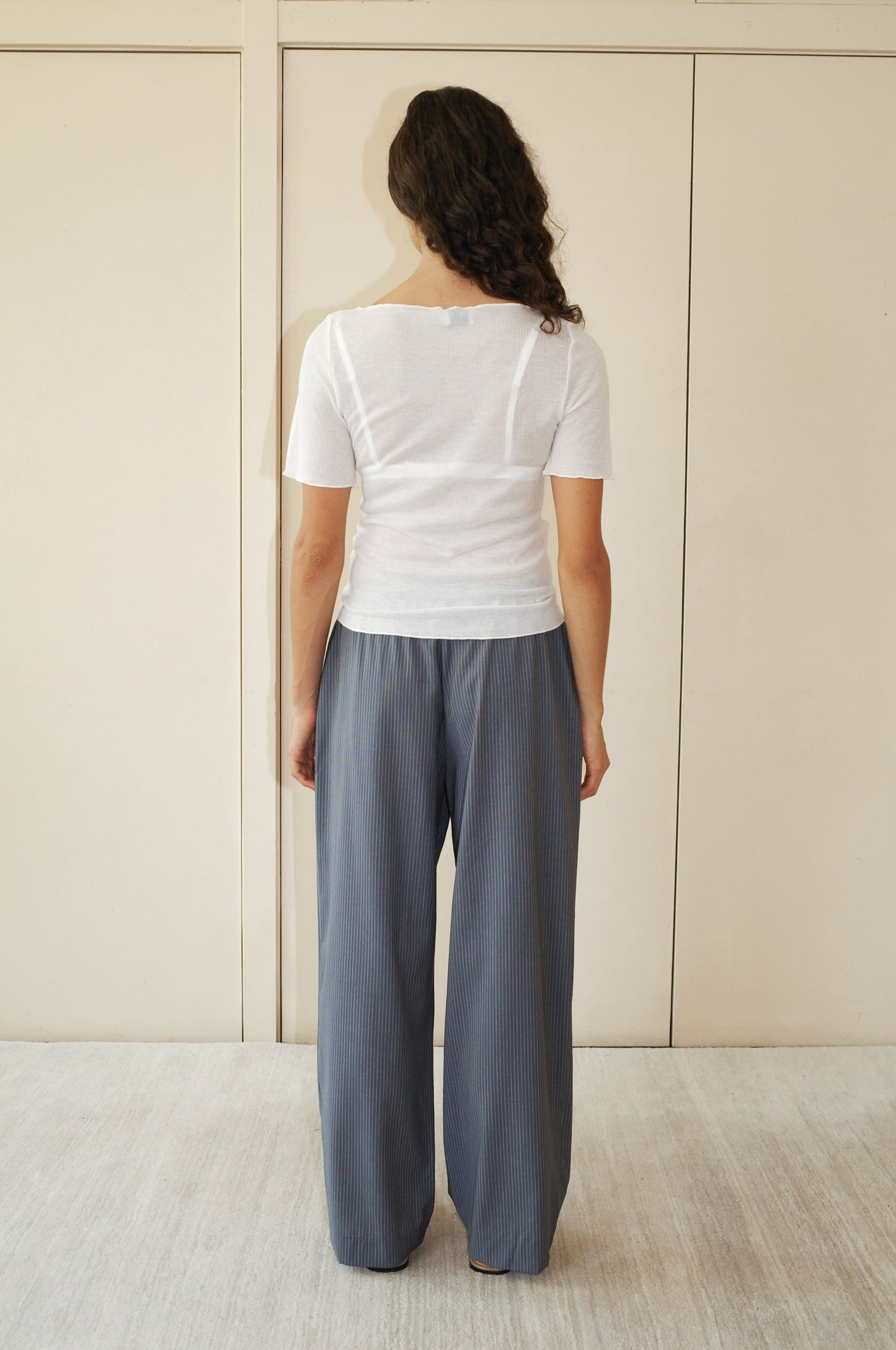 A woman with wavy brown hair faces away from the camera, wearing the Square Knit Tee - Off White with OEKO-TEX 100 certification and loose blue-gray pinstriped pants, standing before a beige wall and light-colored floor.