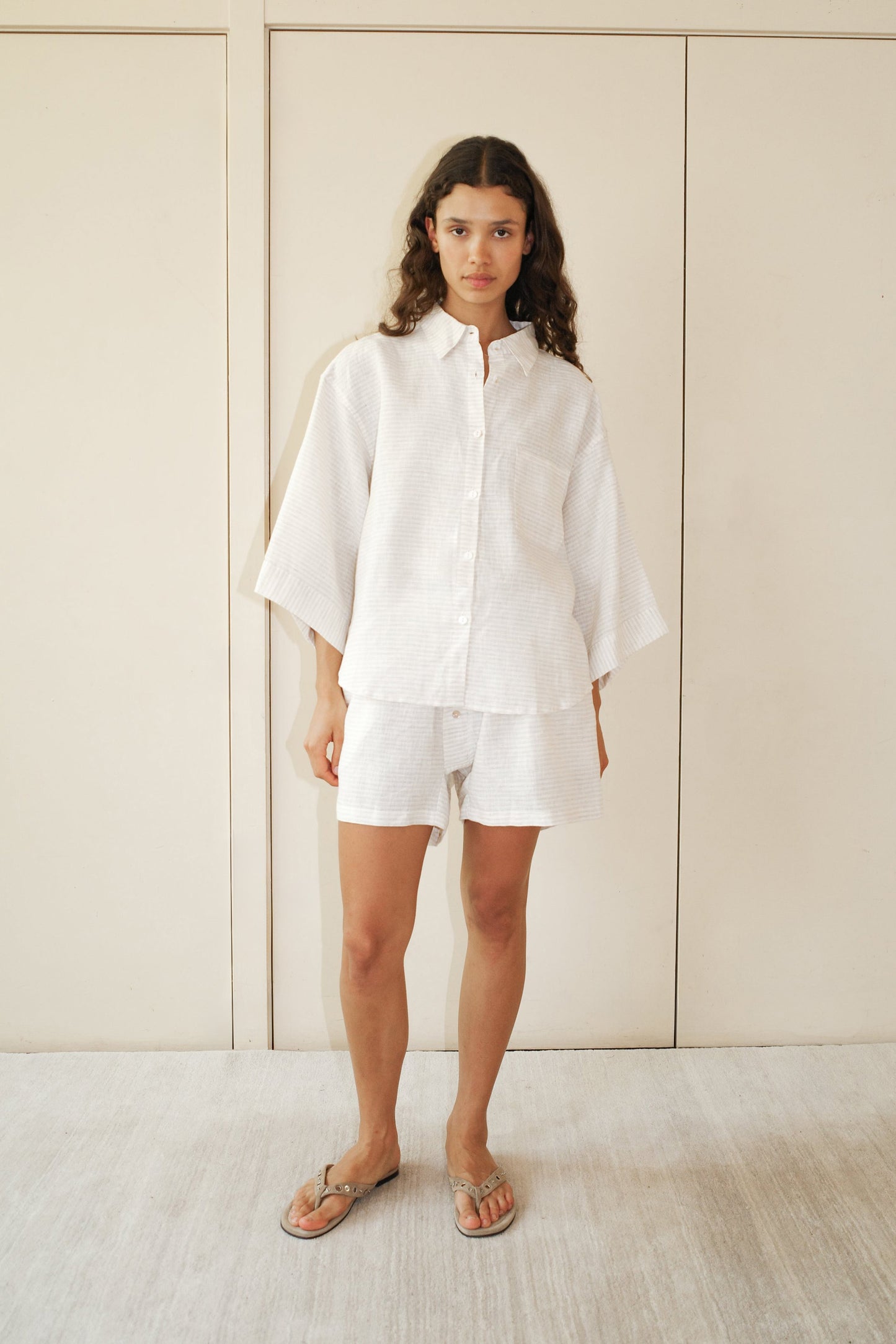 A woman with curly brown hair and a neutral expression stands indoors wearing the 03 Set - Linen Story Stripe, featuring a loose striped linen shirt with wide sleeves and matching boxer-style shorts. She is in front of light-colored cabinets.