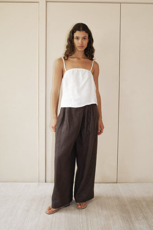 A woman stands indoors against a beige wall, wearing a white sleeveless top, sandals, and the Fold Pant in Slate Check. She has long wavy hair and a neutral expression.
