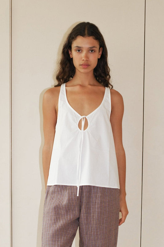 A person with long curly hair wears the Deiji Studios U Shape Top - White, an organic cotton sleeveless top with a front keyhole tie, paired with loose plaid pants, standing against a plain beige background.