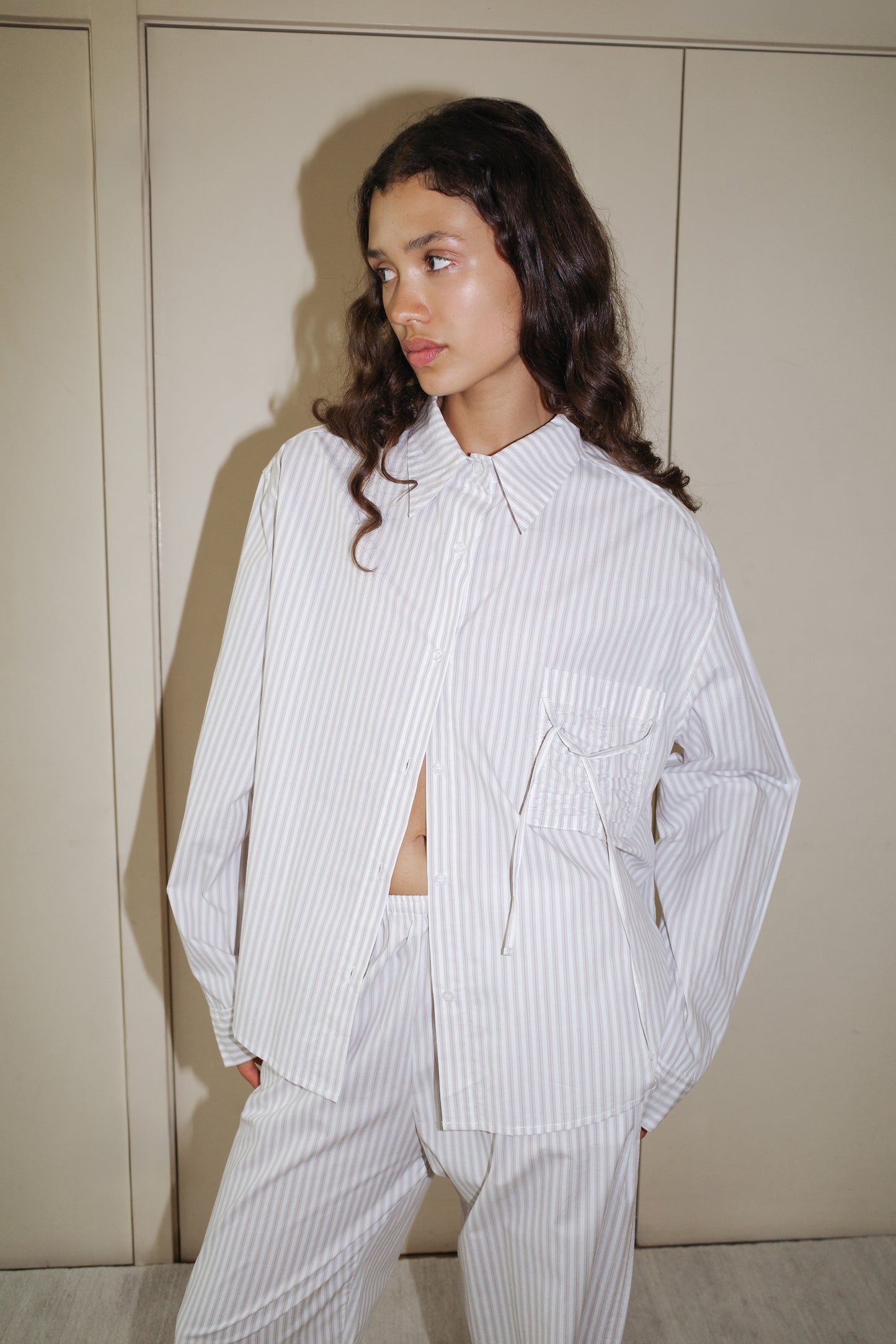 A person with long, wavy brown hair wears Deiji Studios’ Pocket Set - Lake Stripe, a relaxed-fit, light-colored, striped shirt and matching pants in organic cotton poplin, standing indoors by cream doors and looking to the side.