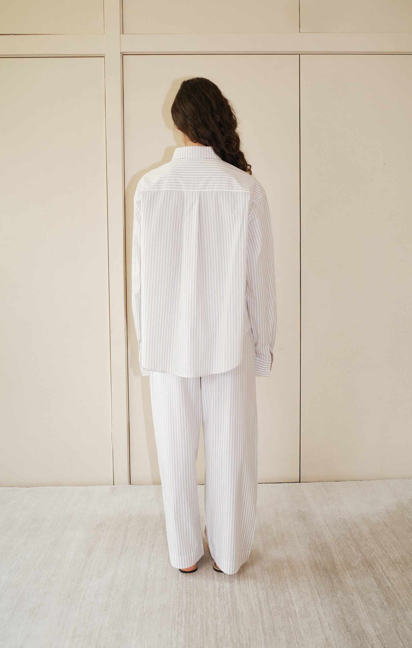 A person stands indoors facing a beige wall, wearing the Pocket Set - Lake Stripe by Deiji Studios: relaxed-fit, white pajama-style pants and a matching long-sleeve, collared shirt with thin vertical stripes. Their hair is long and wavy.