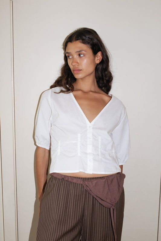 A person with long, wavy dark hair stands against a plain light wall, wearing the Deiji Studios Draw Top - White and brown pinstriped pants with a wrap detail at the waist.