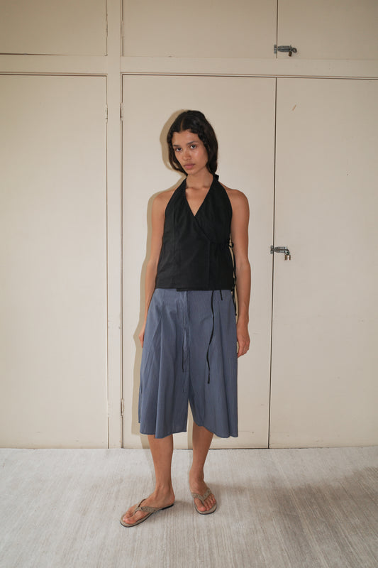 Indoors against a beige wall, a person with short dark hair and a relaxed expression wears the Drawcord Halter, loose blue culottes, and brown flip-flops.