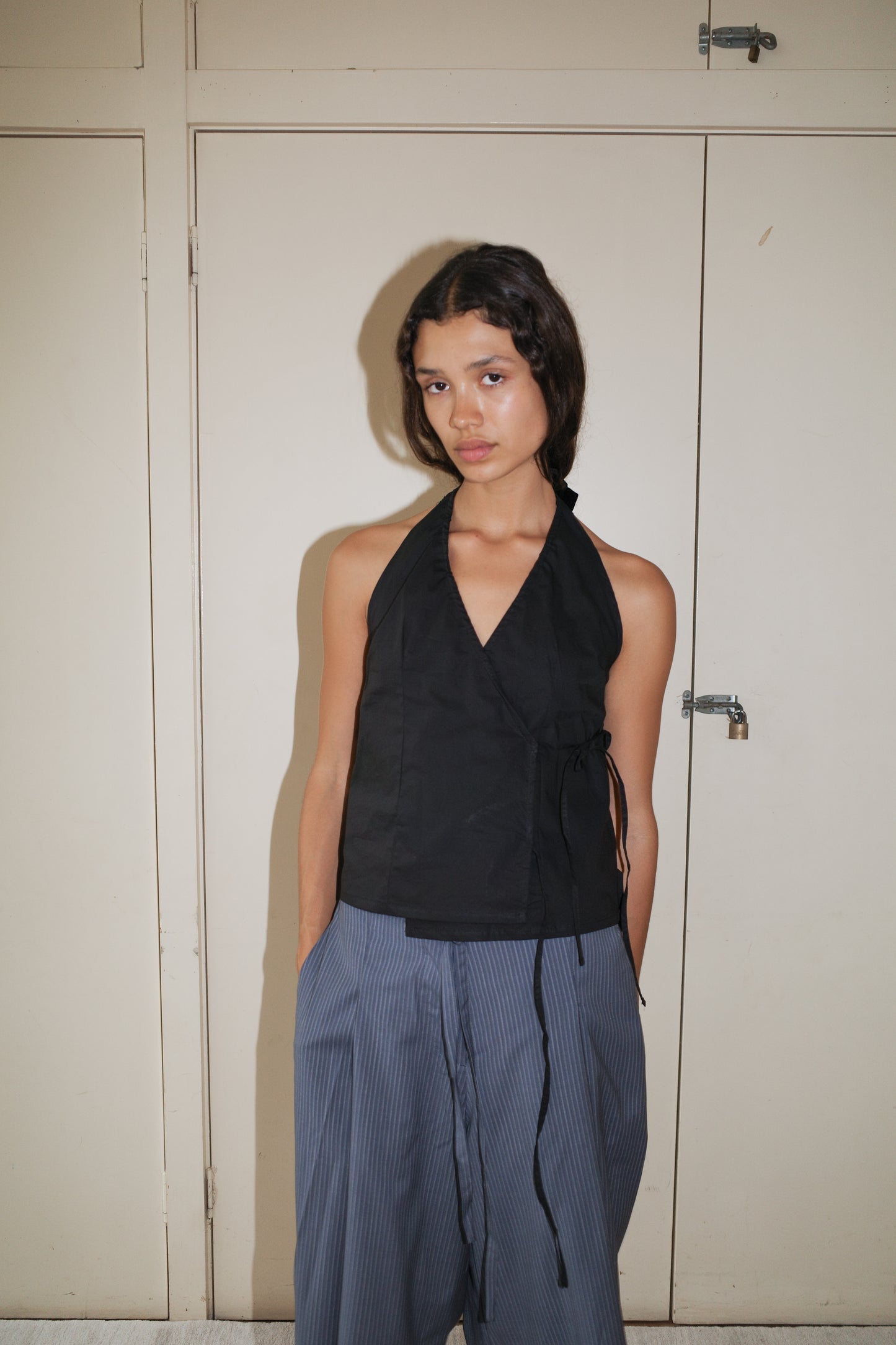 A woman with shoulder-length dark hair and a neutral expression stands against a beige wall, hands in her pockets, wearing the Drawcord Halter—a sleeveless black top—paired with loose blue-gray striped pants.