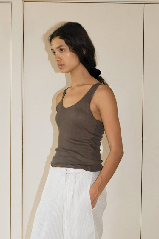 A woman with long dark hair stands indoors against a light wall, wearing the Knit Tank in brown and white pants, her hand in her pocket as she looks thoughtfully to the side.
