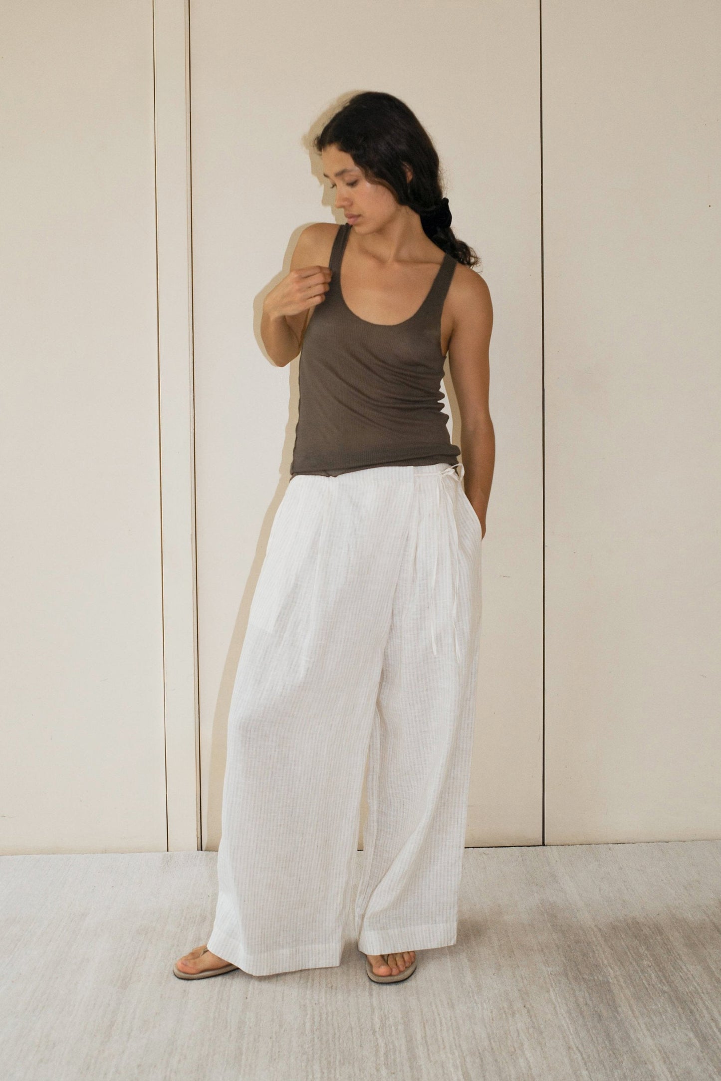 A woman wearing a stone Knit Tank by Deiji Studios, loose white pants, and sandals stands against a beige wall, looking down with one hand tucked in her pocket.