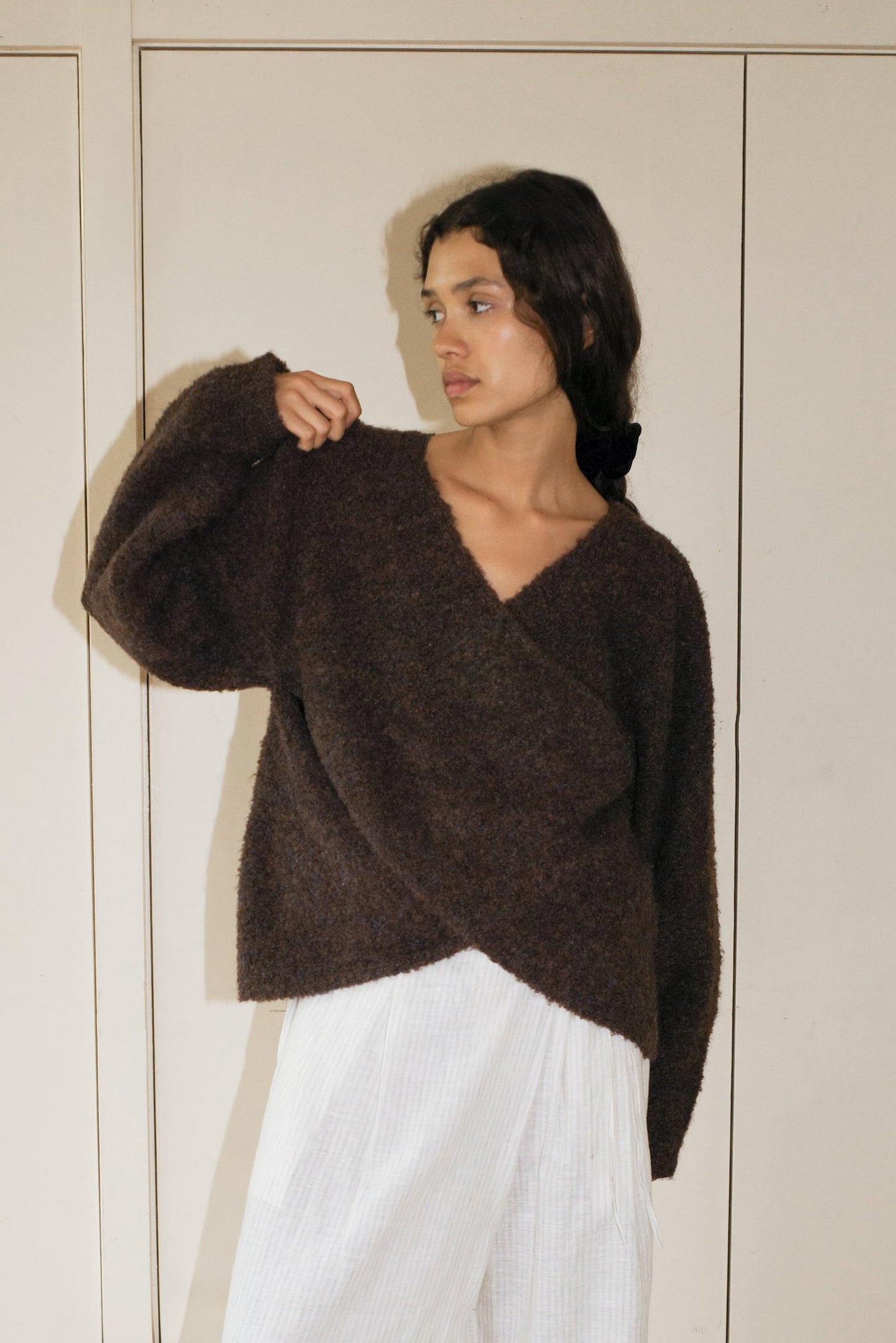 A person stands indoors against a light wall, wearing the Crossover Top - Brown Multi from Deiji Studios with white, loose pants. They look to the side with one arm raised, highlighting this sustainable knitwear piece.
