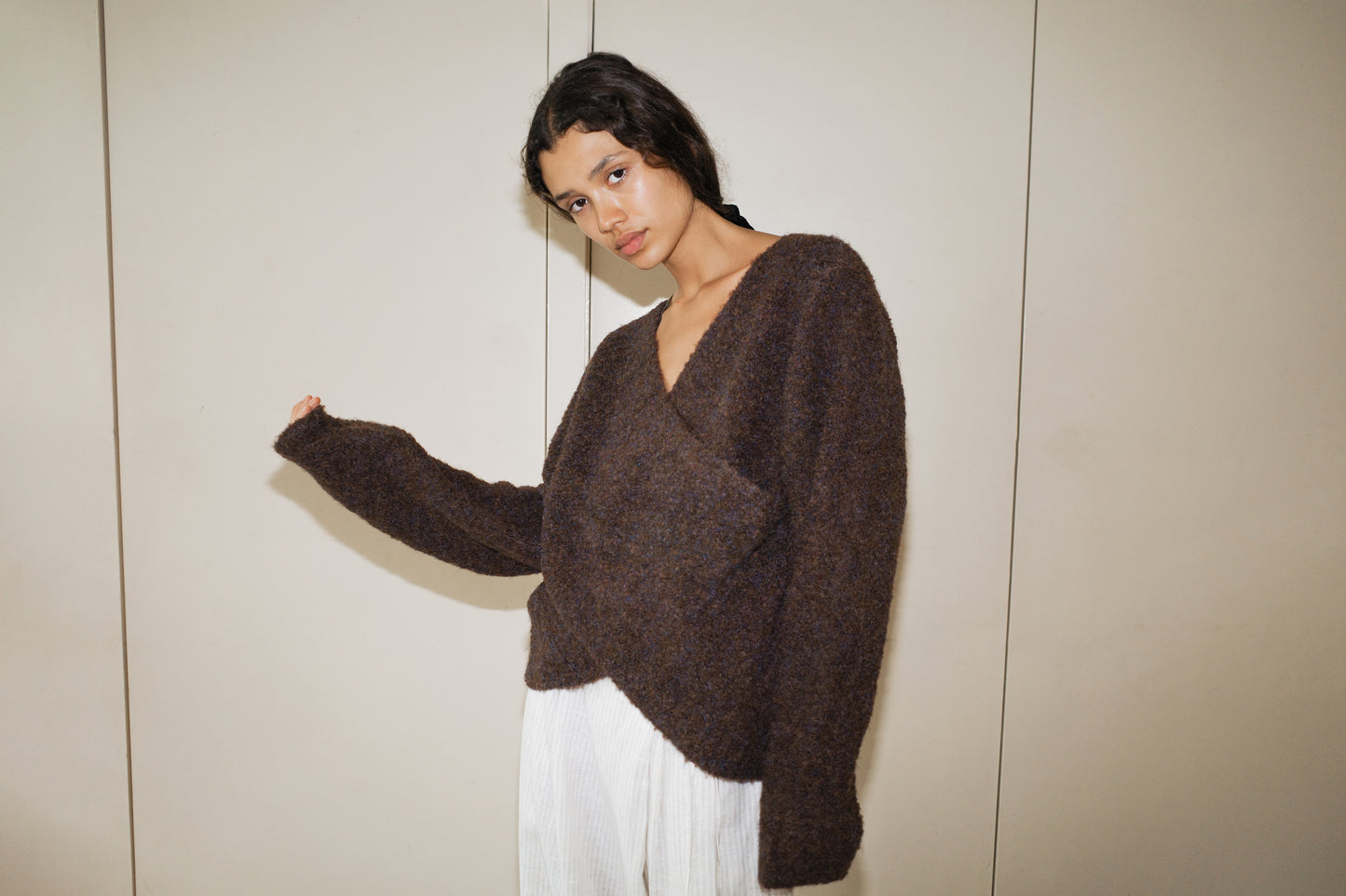 A woman wearing the Crossover Top - Brown Multi, a sustainable knit long-sleeve sweater, and white pleated pants stands against a plain beige wall, gazing at the camera with a relaxed expression.