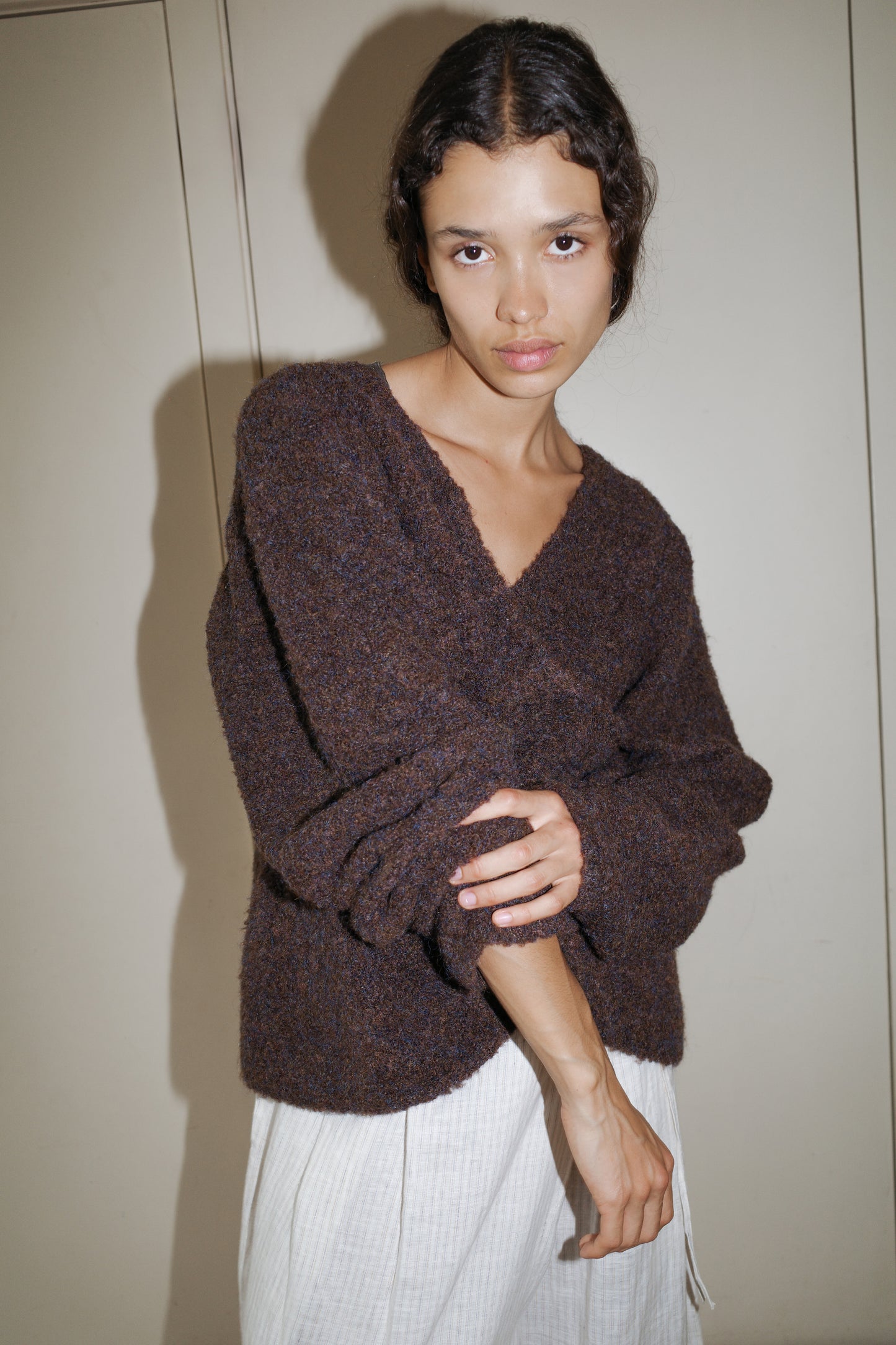 A person with wavy dark hair wears the Crossover Top - Brown Multi and light-colored pants, standing against a beige wall in a relaxed, slightly tilted pose.