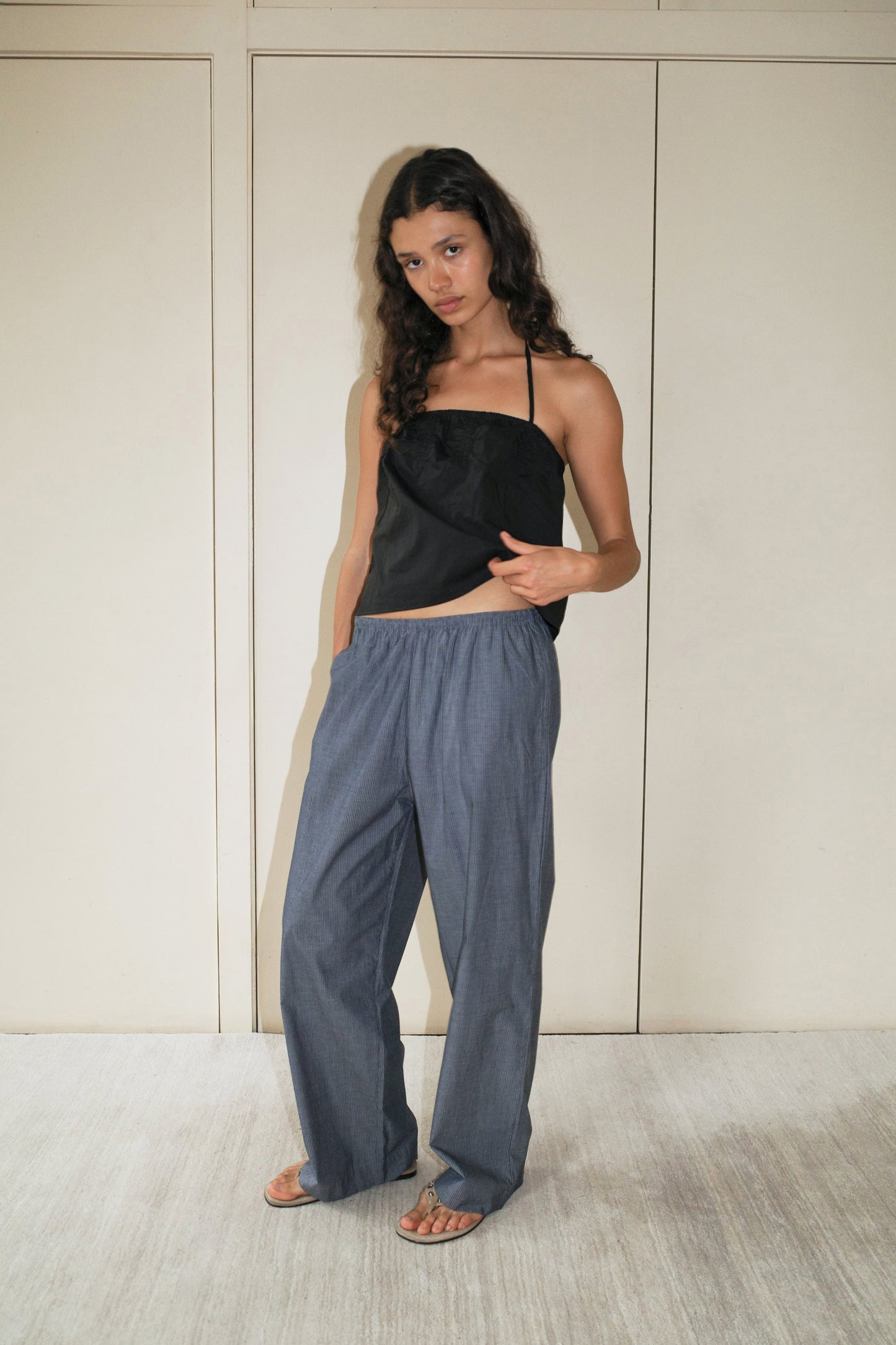 A woman with long curly hair stands indoors against a beige wall, wearing the Ease Trouser in Blue Pinstripe, paired with a black strapless top and sandals, her left hand resting on her top.