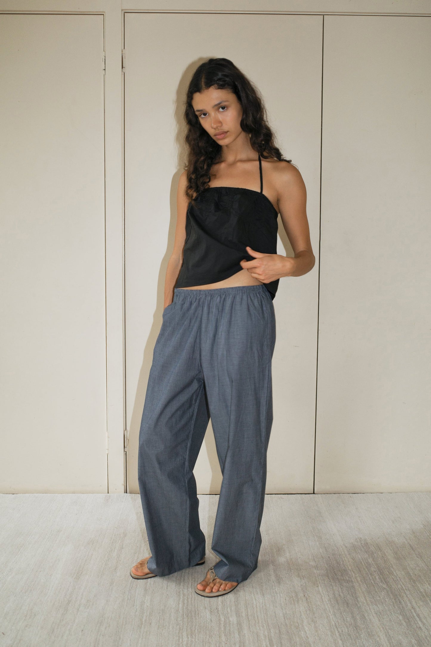 A woman with long curly hair wears a Deiji Studios Tube Top - Black, loose gray pants, and sandals. She stands indoors against beige double doors, looking at the camera in a relaxed pose with one hand holding her top.