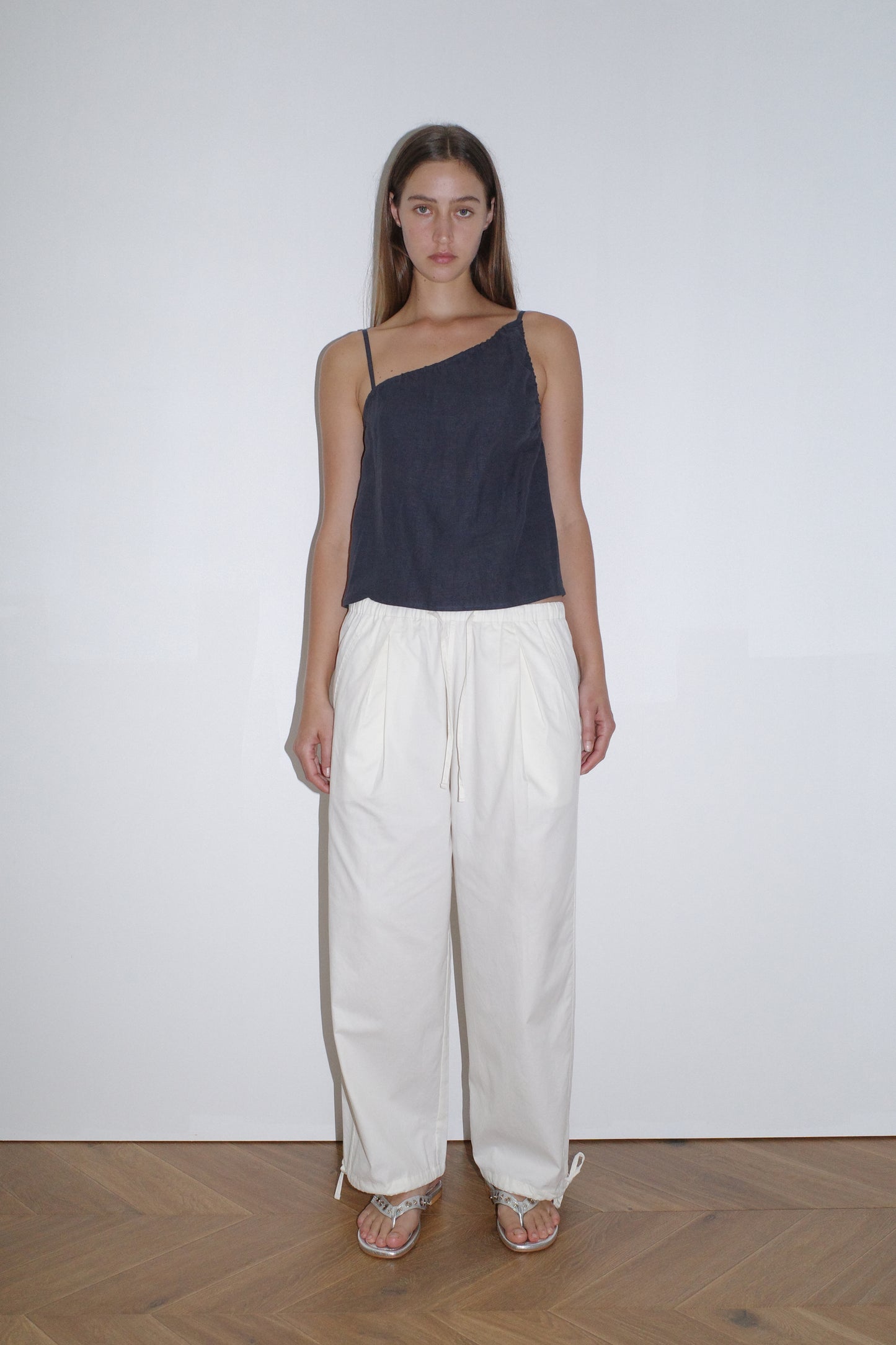 female model wears off white soft cotton trouser featuring a drawcord hem and waist with deep side pockets designed to sit mid-low waist for a relaxed look and made from 100% GOTS certified organic cotton. Paired with the asymmetric tie top in ink, an elevated asymmetric top with a softly gathered front neckline and elastic neckline. Made from 100% GOTS certified organic, OEKO-TEX 100 certified and EU certified stone washed french linen.