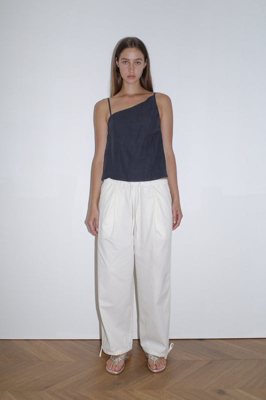 A woman with long brown hair and a neutral expression stands against a plain white wall wearing a Deiji Studios navy one-shoulder sleeveless top, Tie Hem Pant - White, and sandals on light wooden flooring.
