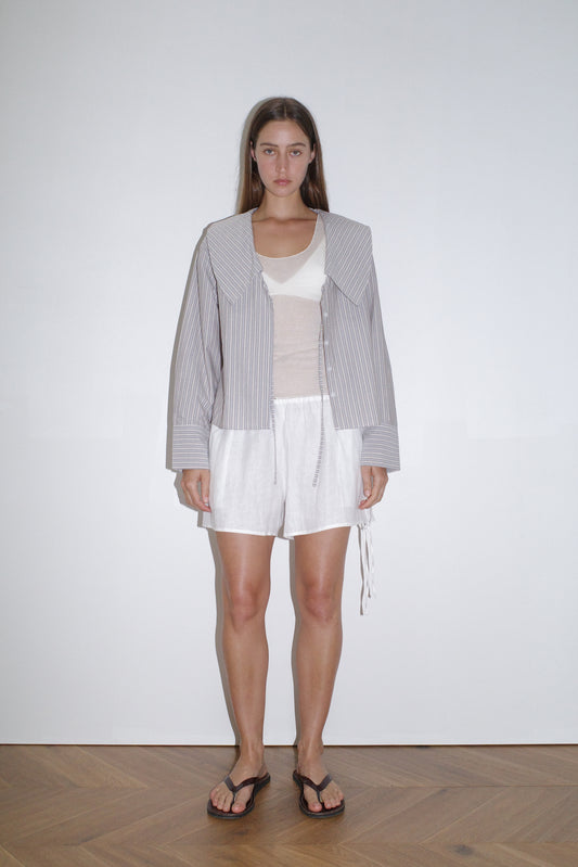 female model wears the collared tie shirt by Deiji Studios in seas stripe
a structured shirt features an oversized collar and cuff, tie at front collar opening, dropped shoulder and elongated sleeves in a boxy fit
made from 100% GOTS certified organic cotton poplin
paired with a knit tank in off white and white linen short
