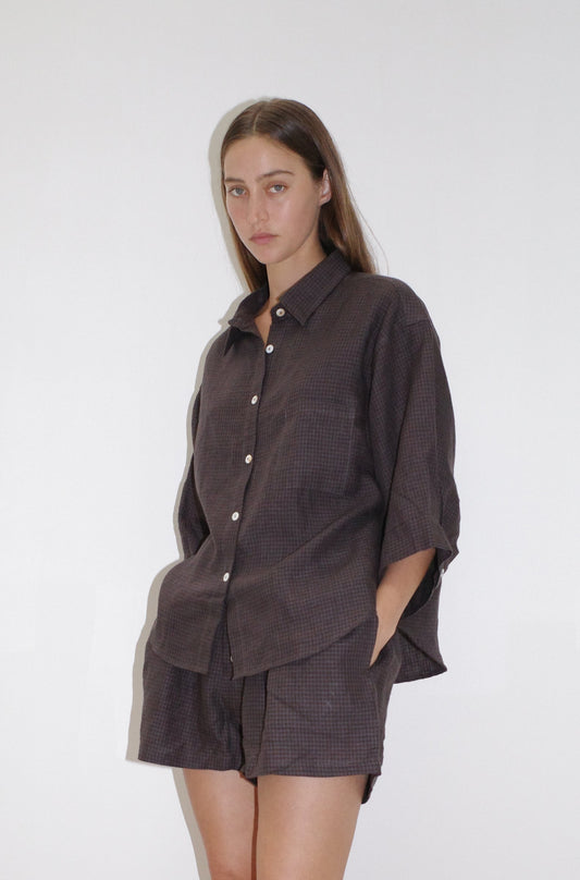A woman with long brown hair and a neutral expression wears the 03 set—loose-fitting, dark plaid button-up shirt and matching shorts—standing against a plain white background with her hands in her shorts pockets.