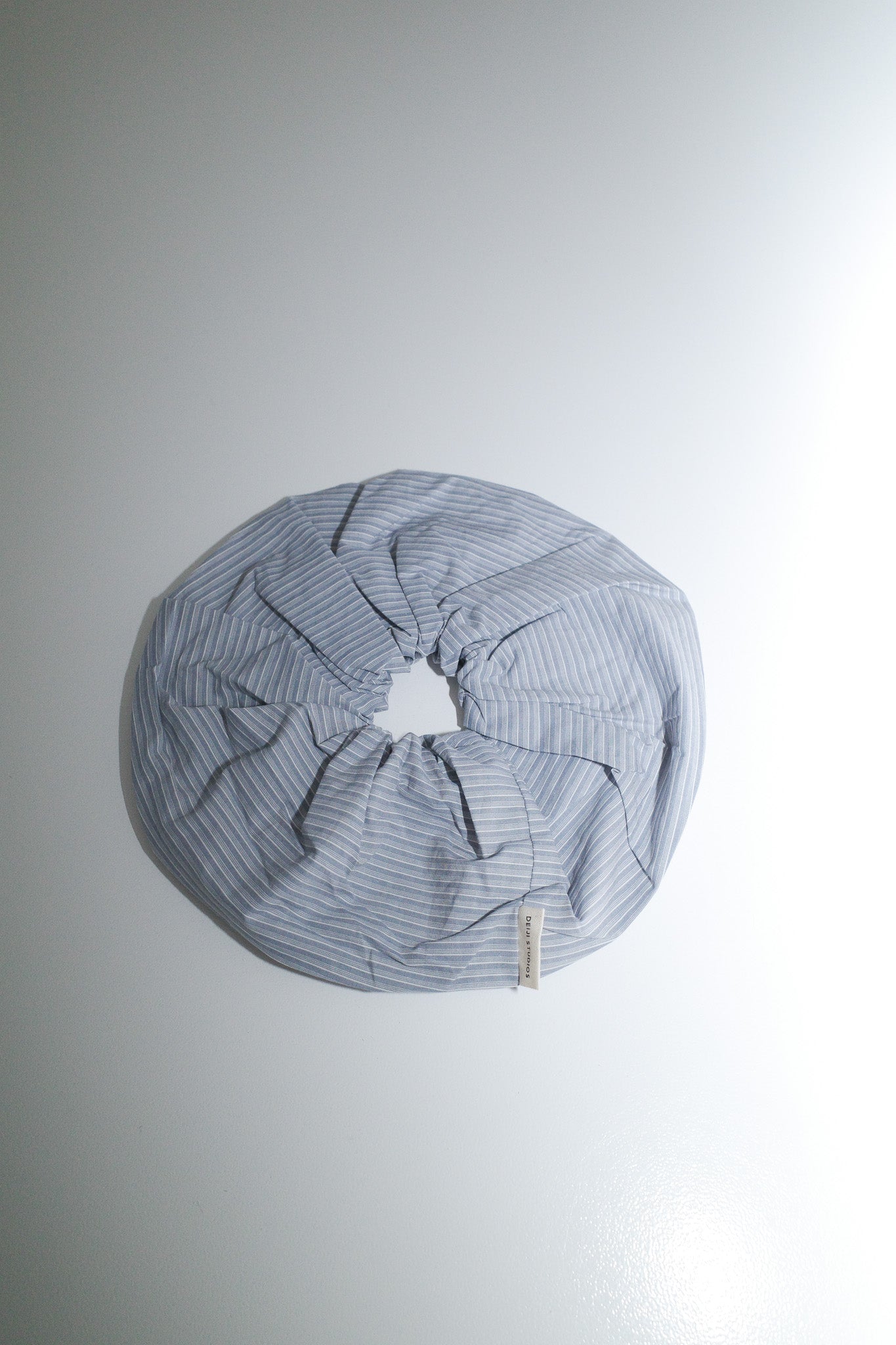 The Scrunchie - Sky Stripe from Deiji Studios, made of oversized organic cotton in light blue and white stripes, is arranged in a circular shape on a plain white surface.