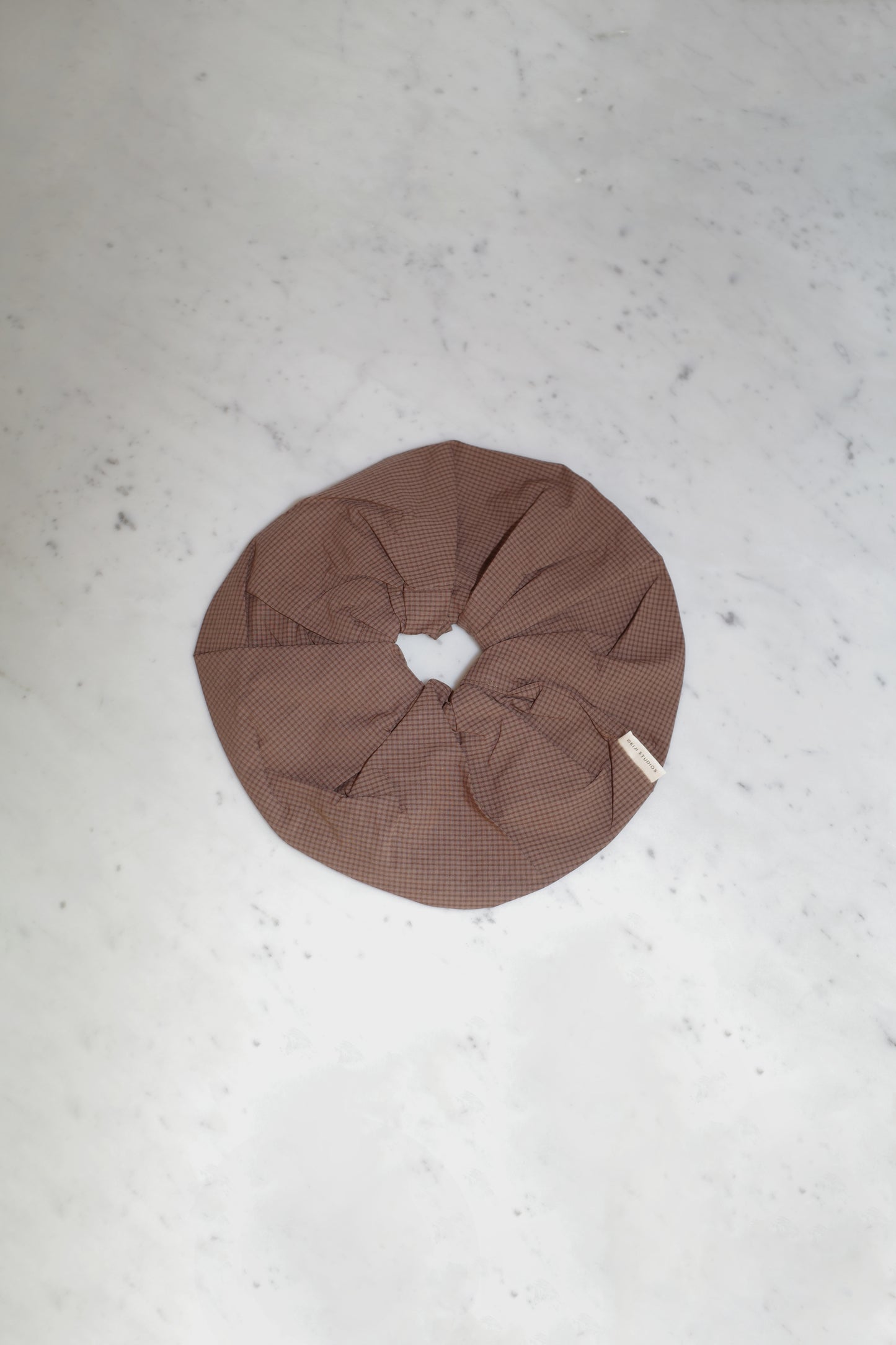 The Scrunchie - Carob Check, an oversized brown organic cotton scrunchie with a subtle checkered pattern from Deiji Studios, rests on a light grey marble surface.