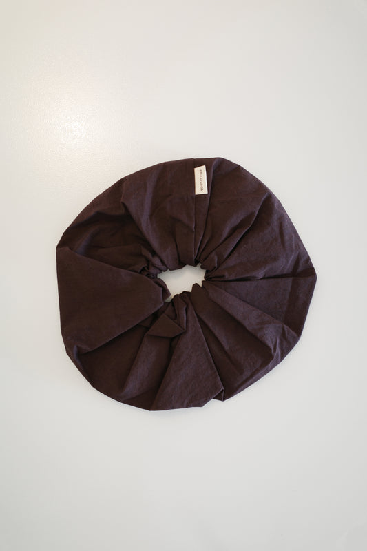 The Scrunchie - Umber from Deiji Studios, a large dark brown oversized scrunchie with a small white tag, is displayed in a circle on a plain white background.