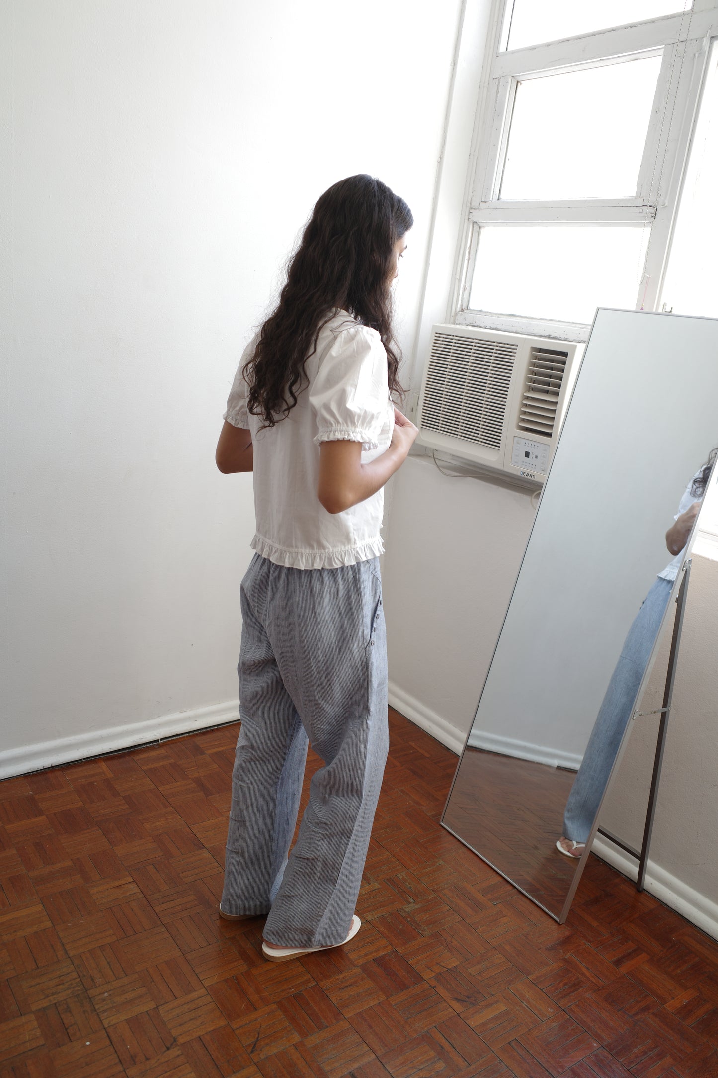A person with long dark hair wearing the Pintuck Top - White from Deiji Studios stands on wooden floors, gazing at their reflection in a tall mirror next to a window with an air conditioner.
