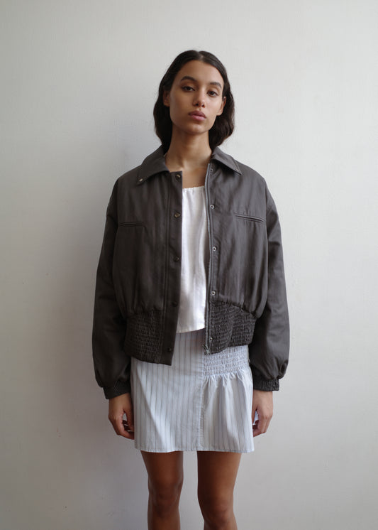 A woman with long, dark hair stands against a white wall, wearing a dark gray Deiji Studios jacket over a white top and the Shirring Skirt - Mineral Stripe. She has a neutral expression.