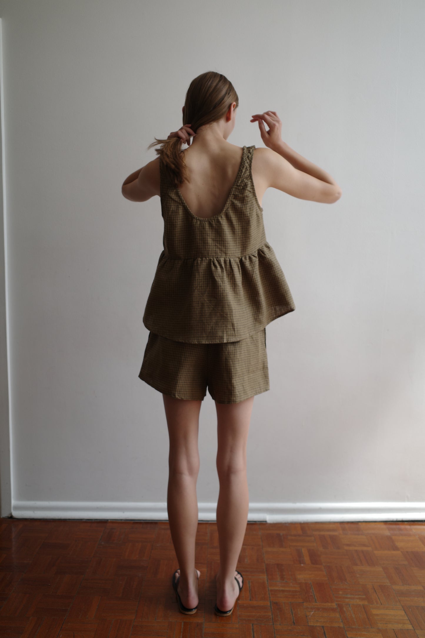 A woman stands indoors on a wooden floor, facing away as she ties her hair. She wears Deiji Studios' Gathered Linen Set - Pine Check, a loose sleeveless top with ruffles and matching shorts, paired with black sandals.