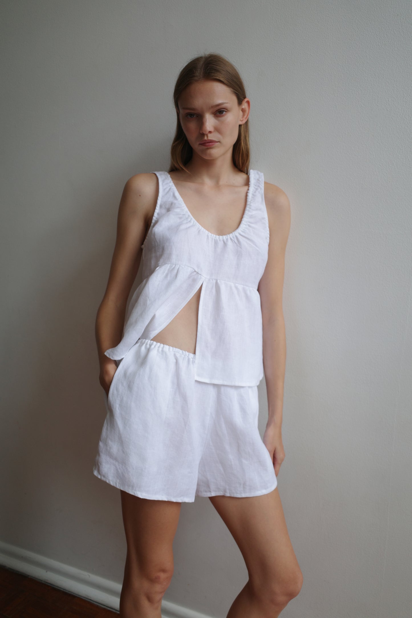 A woman stands against a plain wall, wearing the Gathered Linen Set - White by Deiji Studios—sleeveless, with an asymmetric cutout and matching shorts made from organic French linen—looking directly at the camera with a neutral expression.