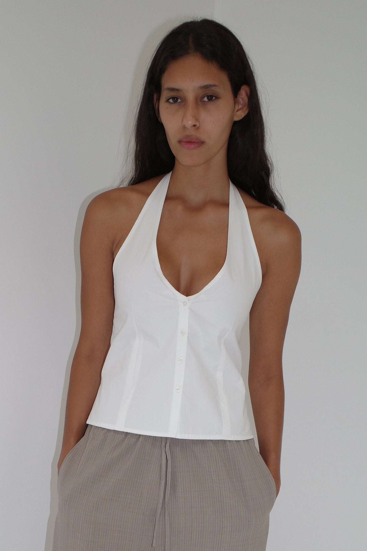 A woman with long dark hair stands against a plain wall, wearing a sleeveless white Waist Halter top and light brown pleated pants, her hands in her pockets and a neutral expression.