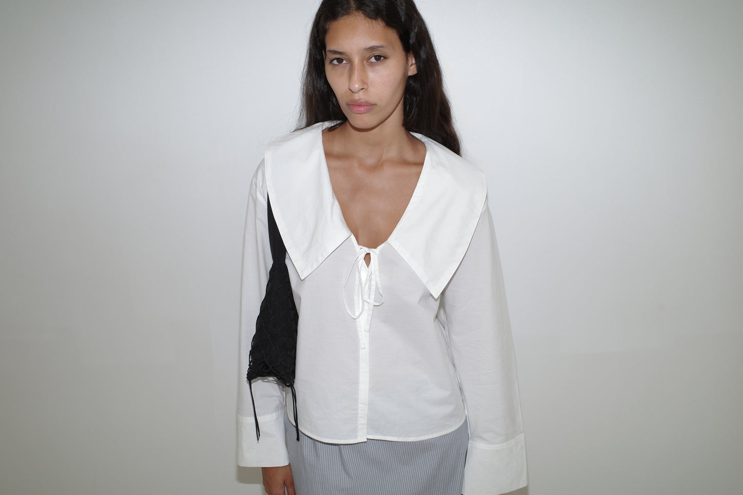A woman with long dark hair wears the Collared Tie Shirt and carries a black bag over her shoulder, standing against a plain white background.