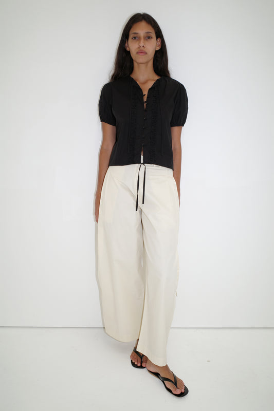 A woman with long dark hair and a neutral expression stands against a plain white background, wearing the Deiji Studios Double Frill Top in black with lace-up detail, wide-leg white pants, and black flip-flops.