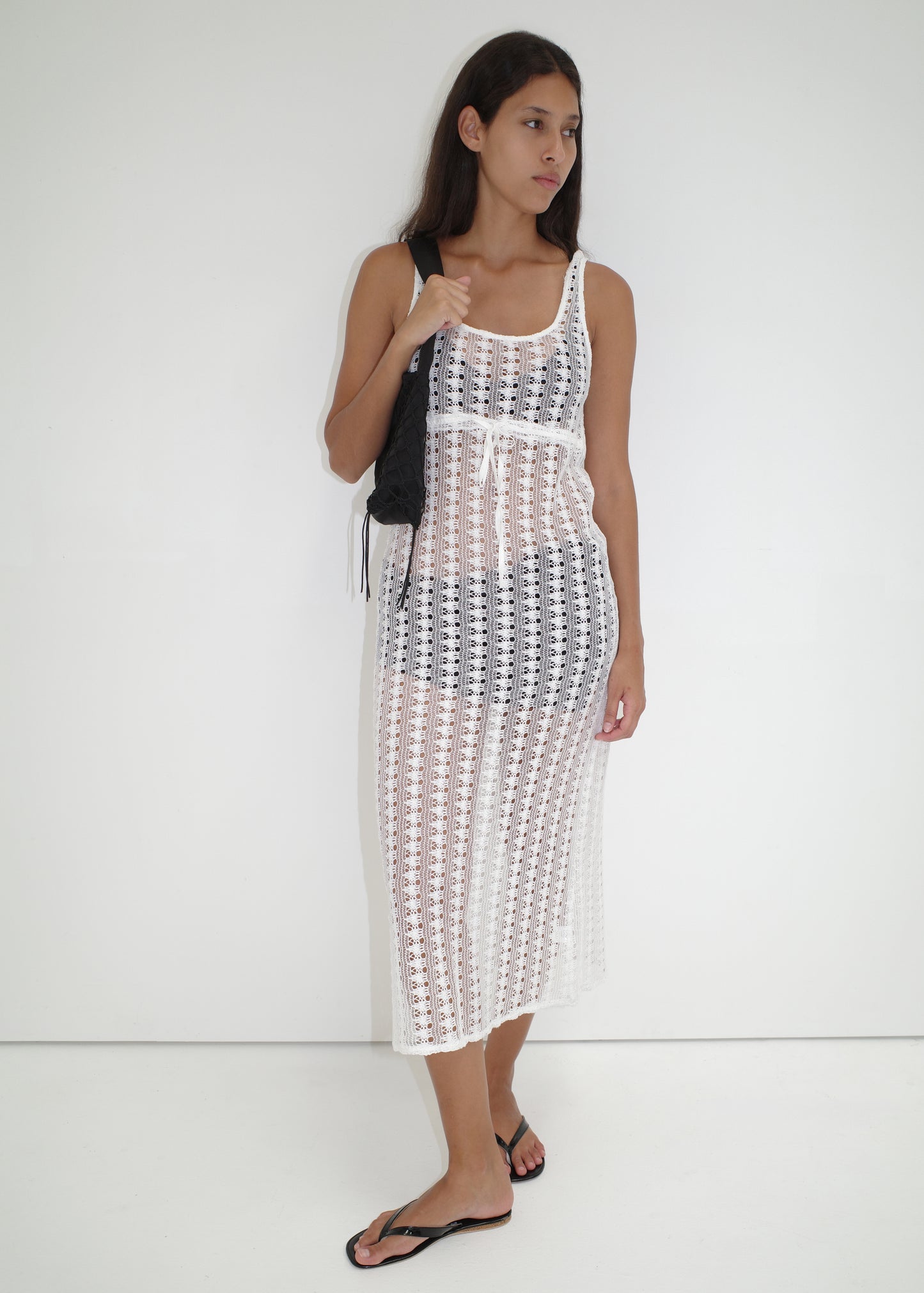 A woman stands against a white background wearing the Thread Dress - Off White by Deiji Studios over a black bikini, paired with black flip-flops and a black shoulder bag. She looks to the side with a relaxed expression.