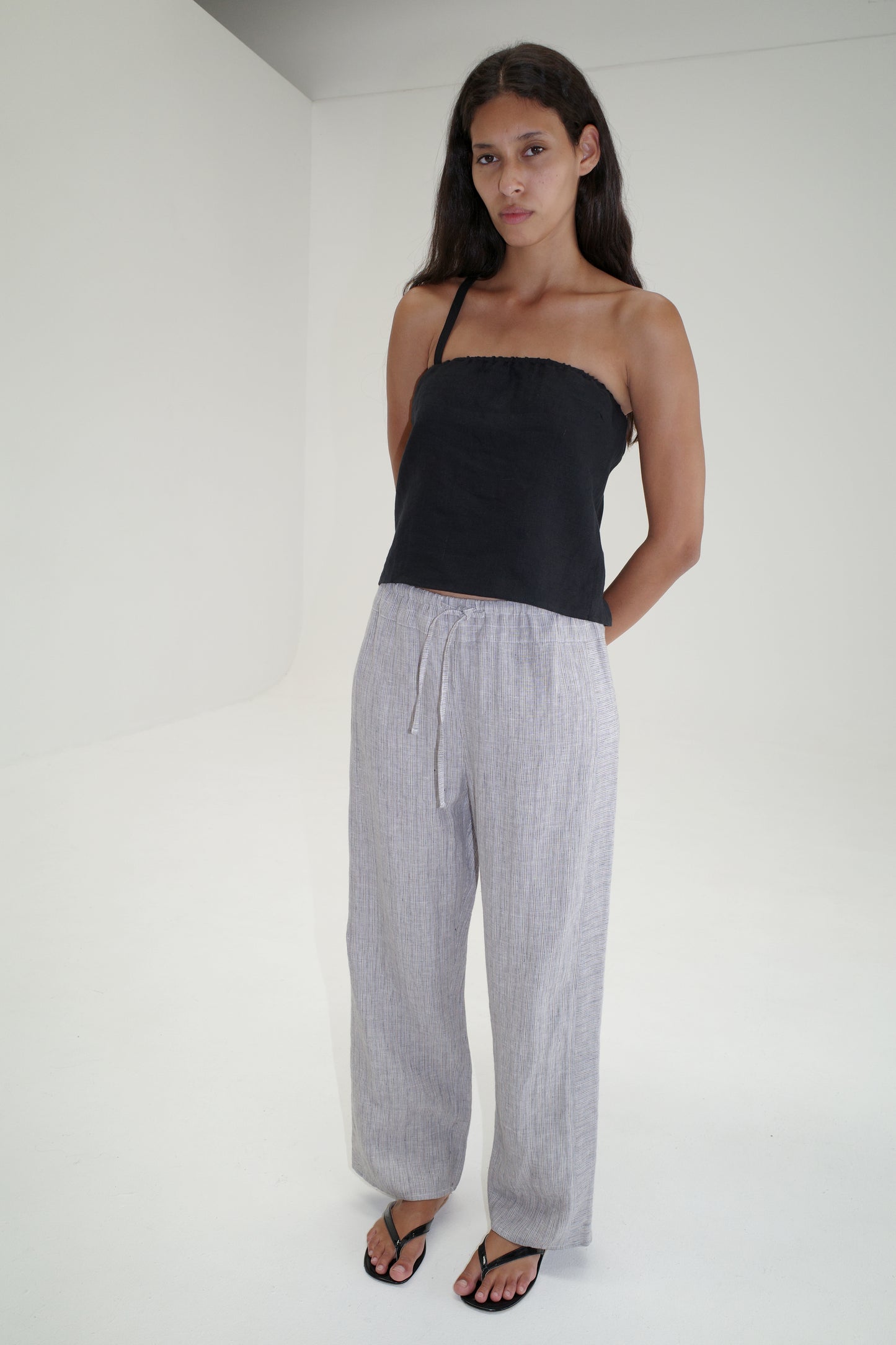 A person with long brown hair stands in a minimal white room, wearing a black strapless top, Double Waist Linen Pant - Haze Blue, and black sandals. Their arms are relaxed behind their back.
