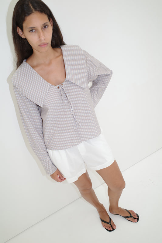 A woman with long dark hair wears the Collared Tie Shirt, featuring light stripes and a large collar, paired with white shorts and black flip-flops, as she stands against a white wall looking slightly up at the camera.