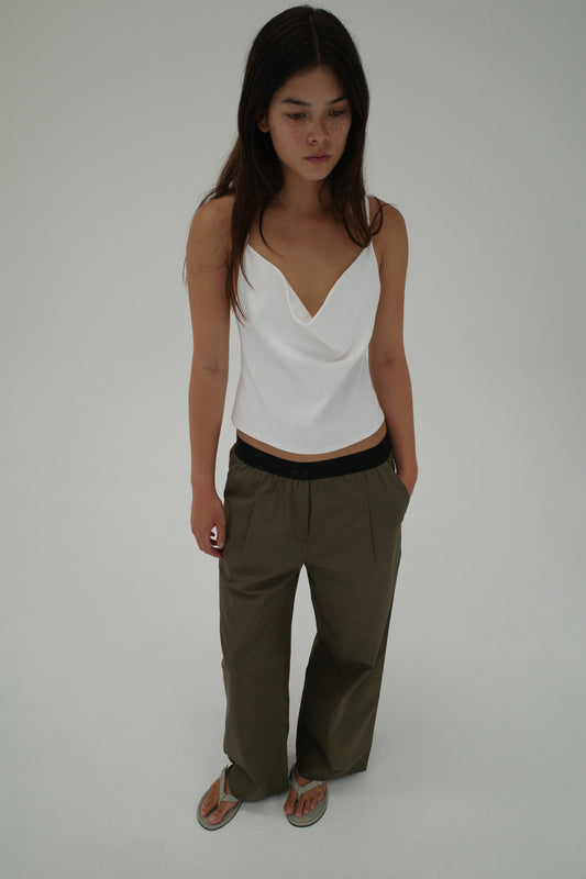 A woman with long dark hair wears a white sleeveless top, Deiji Studios' Contrast Waist Pant - Olive, and sandals as she stands against a plain white background.
