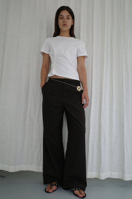 A model stands against a white draped backdrop wearing a fitted white short-sleeve t-shirt, Bind Pant - Charred Black Stripe by Deiji Studios with chain and disc waist details, and black embellished flip-flops.