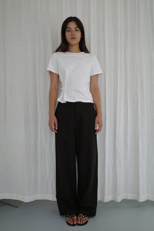 A woman with long dark hair stands in front of white curtains, wearing the Pinch Tee - White by Deiji Studios, loose black pants, and black sandals. She has a neutral expression and her arms are relaxed at her sides.