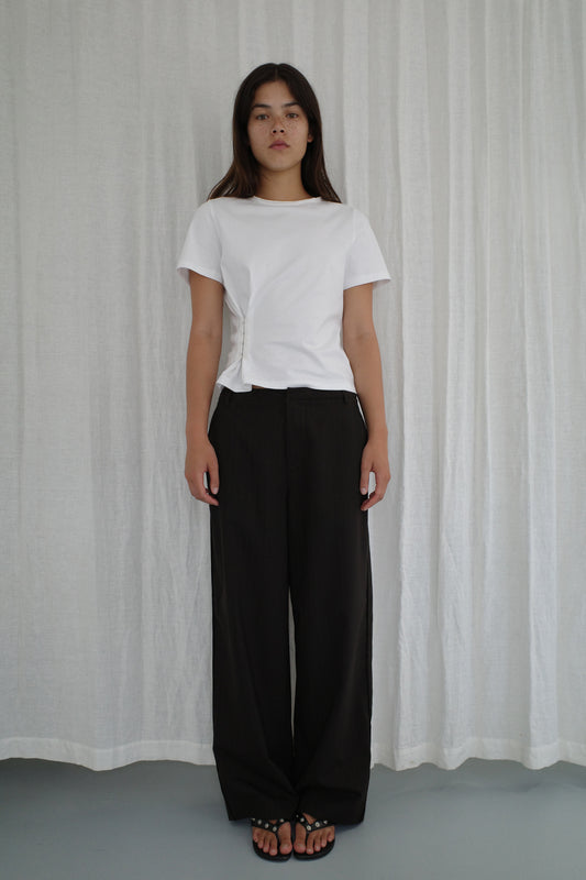 A woman stands before a white curtain, wearing a white short-sleeve t-shirt, the Bind Pant in Charred Black Stripe by Deiji Studios, and black sandals. She has a neutral expression with her arms relaxed at her sides.