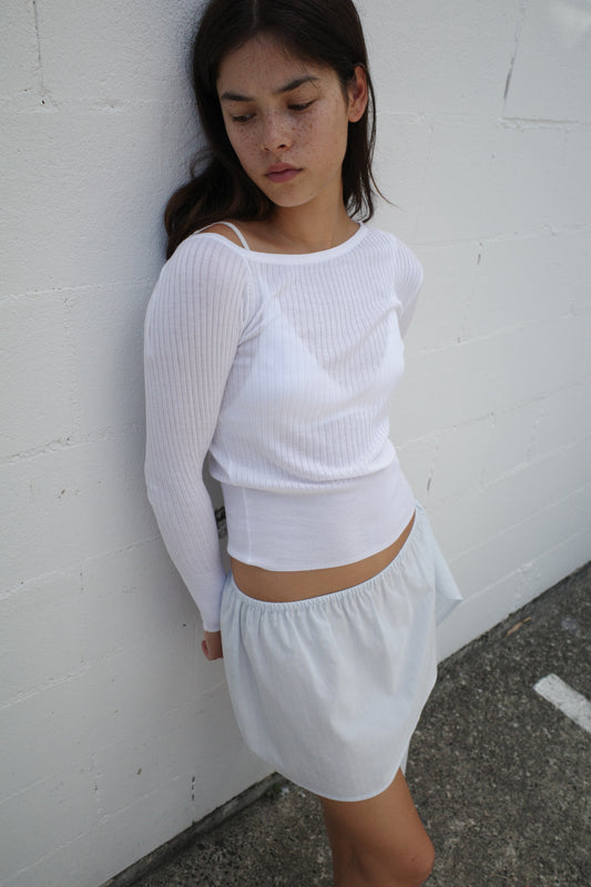 A woman with long dark hair stands against a white textured wall, wearing the Shoulder Knit Top - White and a light, high-waisted skirt. She gazes downward with a pensive expression.