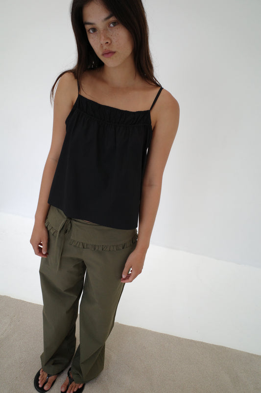 A woman with long brown hair wears a loose black spaghetti strap top, the Flounce Belt Pant in khaki, and black sandals, standing indoors against a plain white wall and beige floor, gazing softly at the camera.