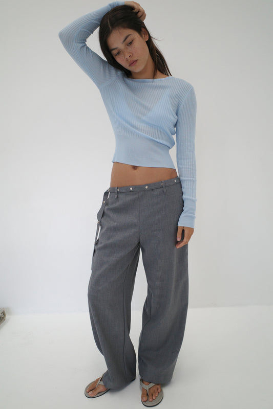A woman stands against a white background wearing the Shoulder Knit Top - Blue from Deiji Studios and loose gray belted pants. Barefoot, she raises one arm to touch her head and gazes downward.