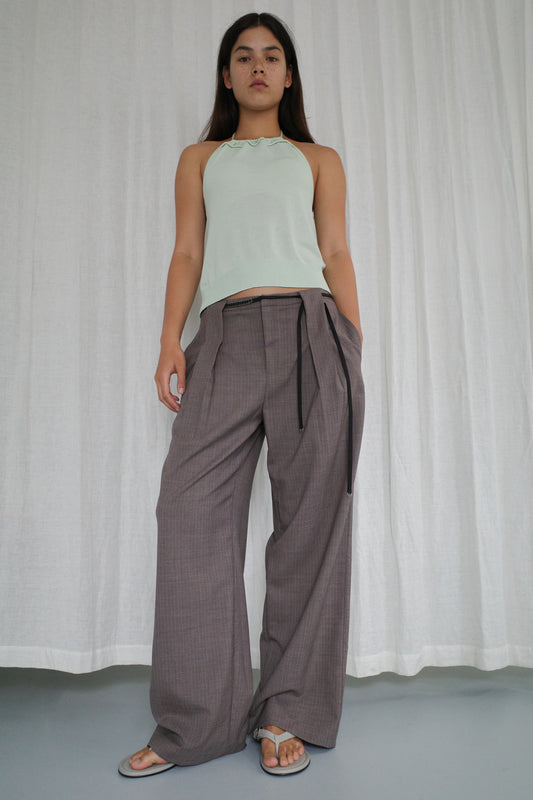 A woman stands in front of white curtains wearing a light green halter top, Belt Loop Pant - Grey Pink Stripe, and gray sandals. She looks confidently at the camera.