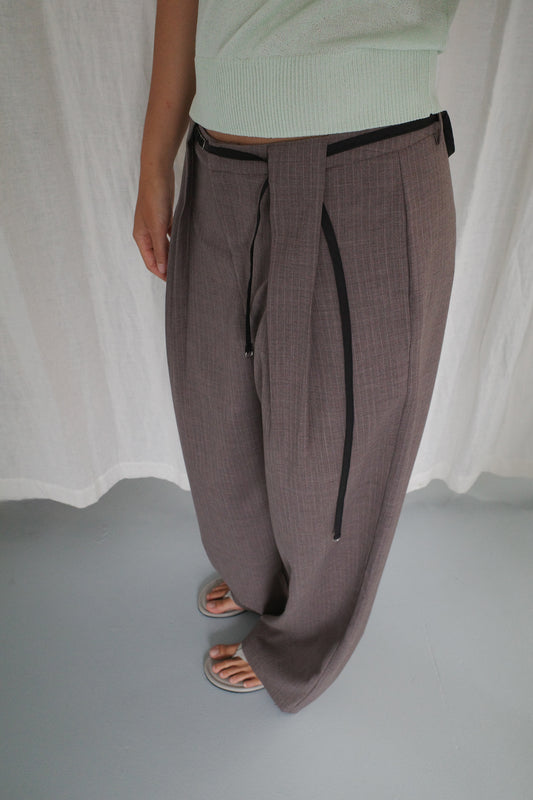 A person stands indoors wearing Belt Loop Pant - Grey Pink Stripe, paired with a light green top and gray open-toe sandals, against a backdrop of a white curtain and light gray floor.