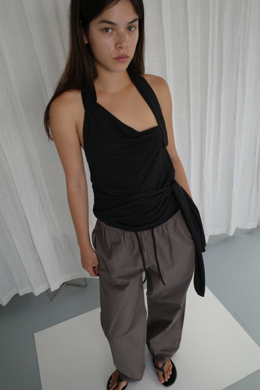 A woman with long dark hair stands indoors in front of white curtains, wearing a sleeveless black halter top, Layered Pant - Khaki Check, and black sandals. She looks slightly up at the camera with a neutral expression.