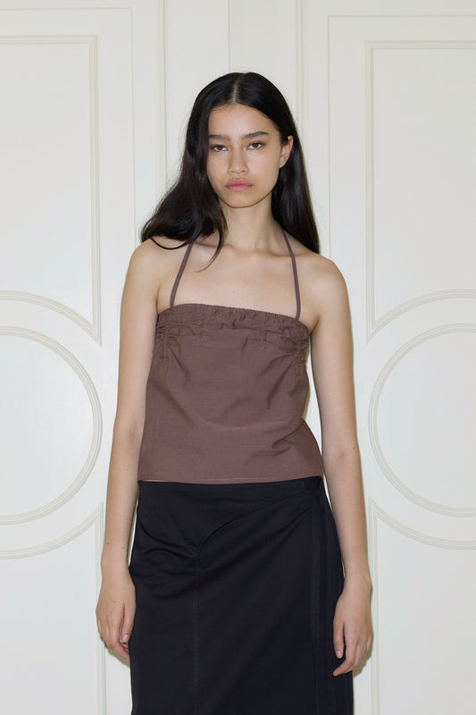A woman with long dark hair wears a brown tube top and a black skirt, standing in front of cream-colored double doors. She has a neutral expression and relaxed arms at her sides.