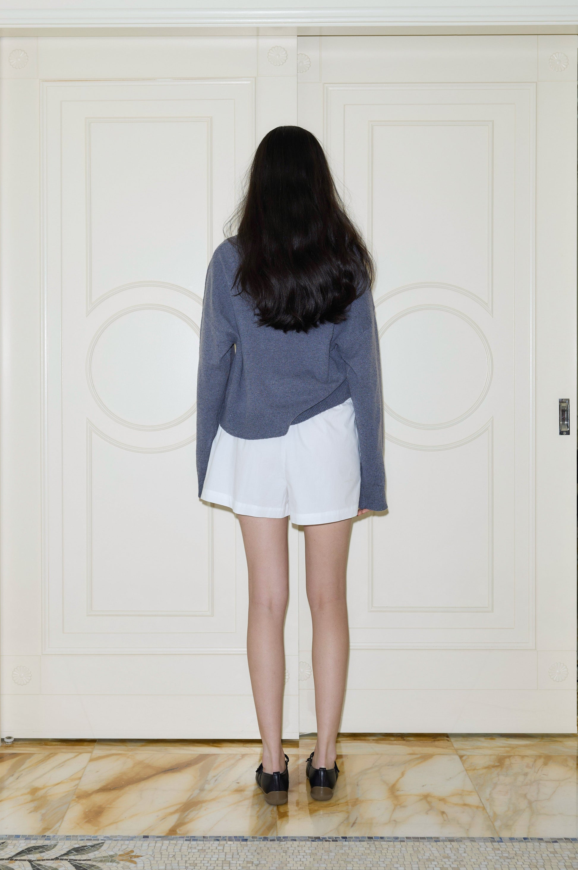 A person with long, dark hair, dressed in a blue long-sleeve shirt, black shoes, and the Organic Cotton Boxer - White, stands on a marble floor facing a closed white double door. The individual is viewed from behind.