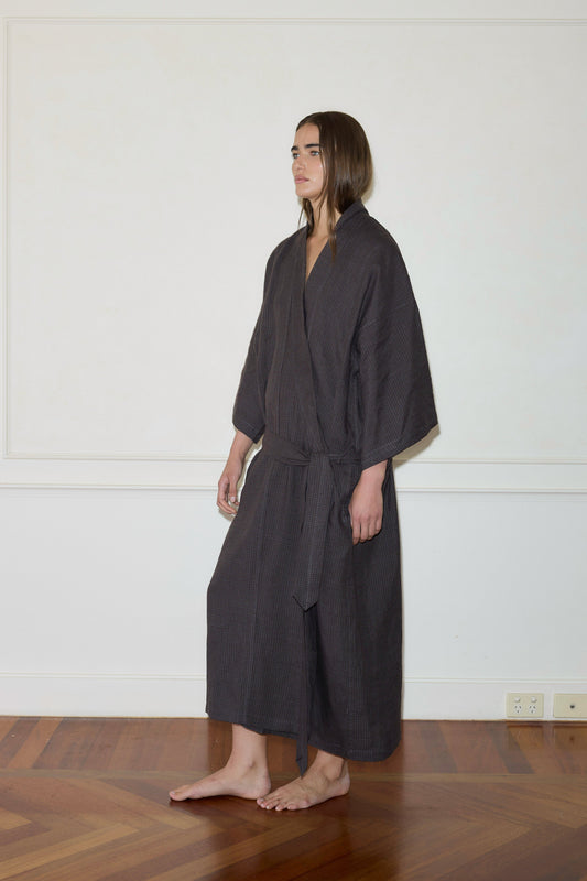 A person with long hair stands barefoot on a wooden floor, wearing the 02 Robe - Slate Check from Deiji Studios, tied at the waist. The background features a light-colored wall with white trim.