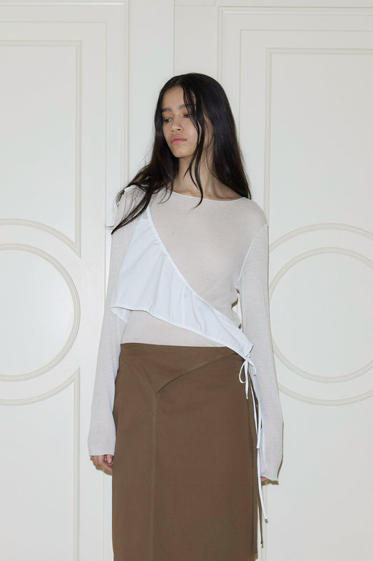 A woman with long dark hair stands against a white paneled door, wearing a sheer beige top, brown skirt, and the Cotton Sash - White by Deiji Studios styled asymmetrically across her chest. She gazes downward with a neutral expression.